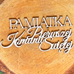 Scrapka z napisem „Pamiątka Pierwszej Komunii Świętej” z białej tektury, ułożona na tle plastra drewna.