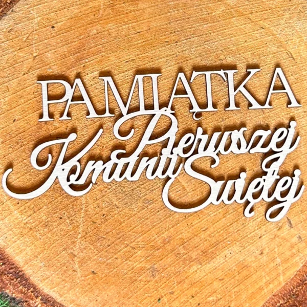 Scrapka z napisem „Pamiątka Pierwszej Komunii Świętej” z białej tektury, ułożona na tle plastra drewna.